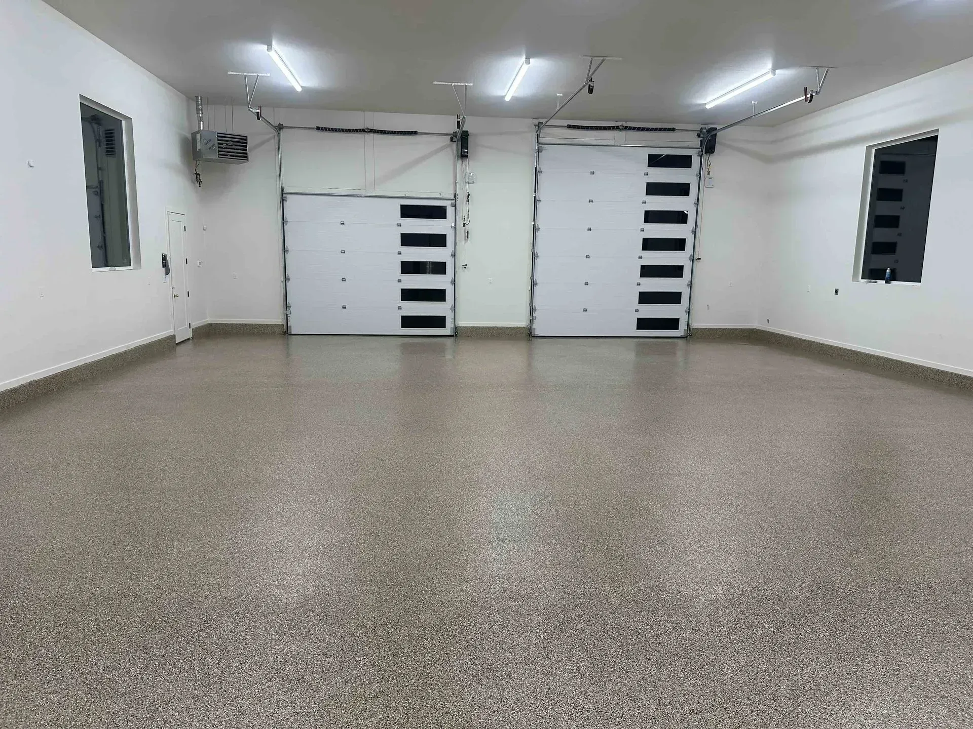 An empty garage with a grey speckled epoxy floor, white walls, two closed white garage doors, and bright ceiling lights.