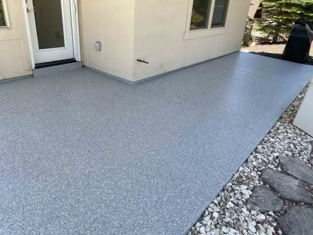 A light gray speckled epoxy-coated concrete patio sits adjacent to a white house wall and a stone garden bed.