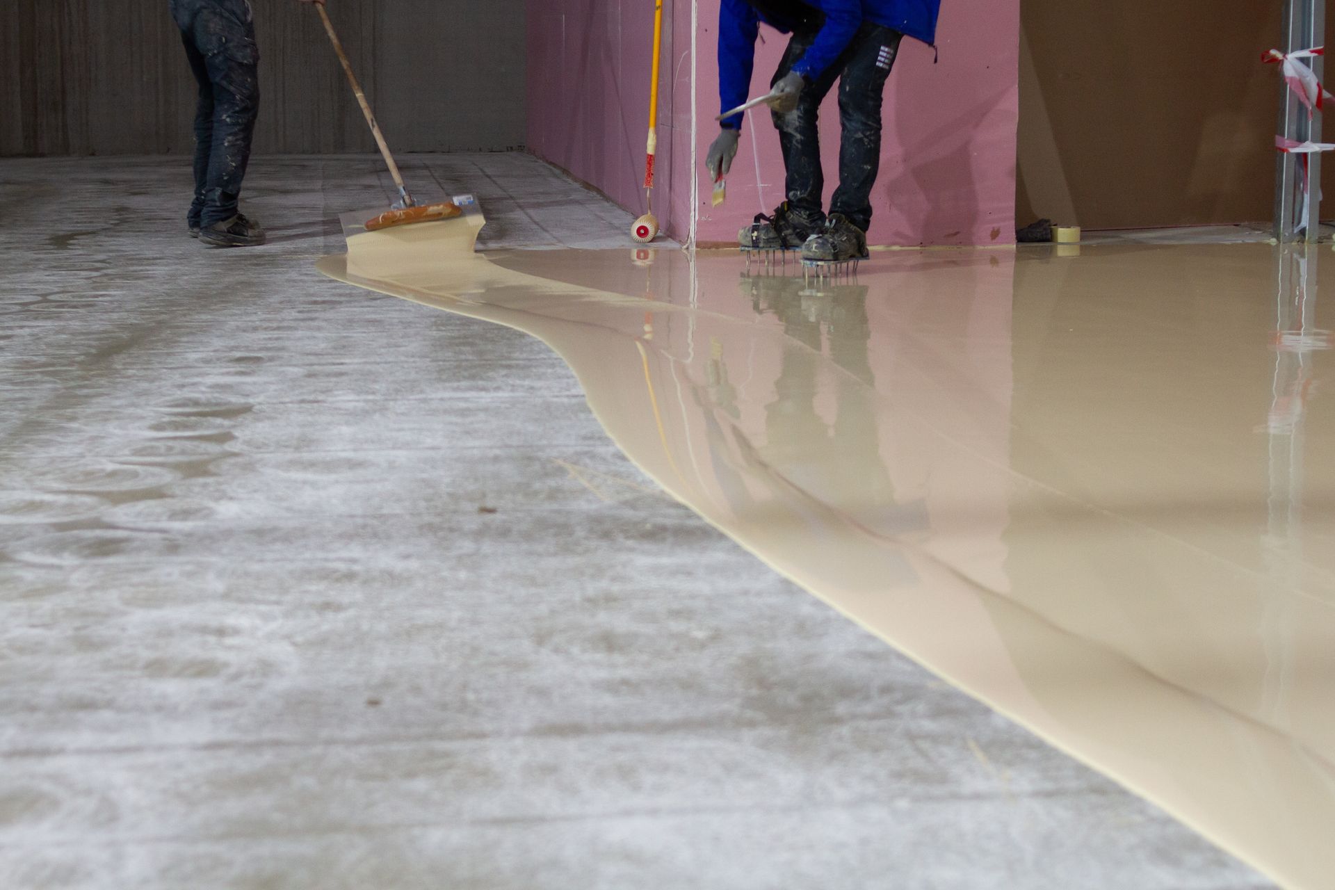 Workers spreading beige epoxy coating over a gray concrete floor in an indoor space.