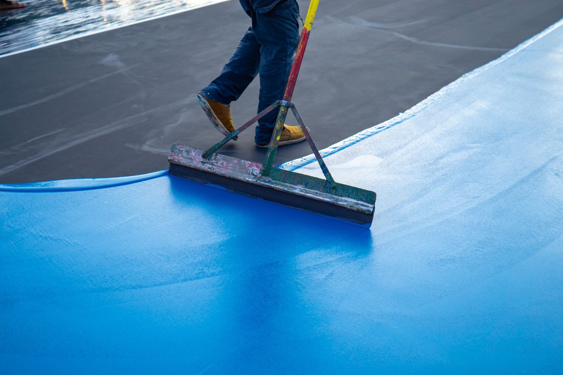 A person uses a squeegee to apply bright blue coating to a dark surface.