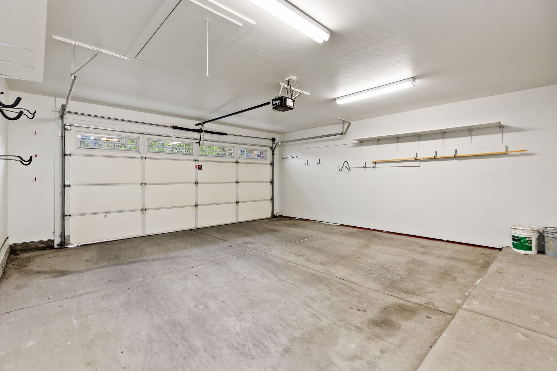 A spacious, empty two-car garage with white walls, a concrete floor, overhead lighting, and a white paneled garage door.