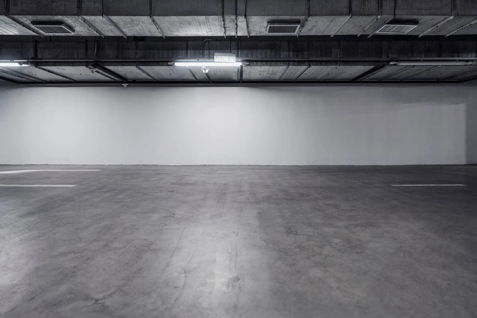 A vast, empty underground concrete parking garage with a long white wall and industrial overhead lighting.