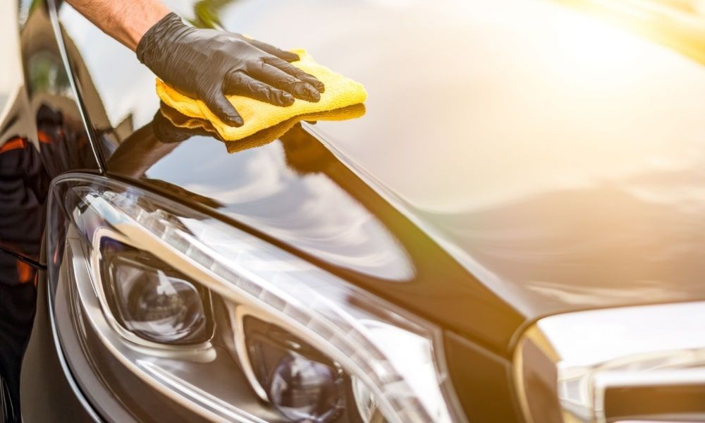 A gloved hand wipes a yellow cloth across a shiny black car hood, cleaning.