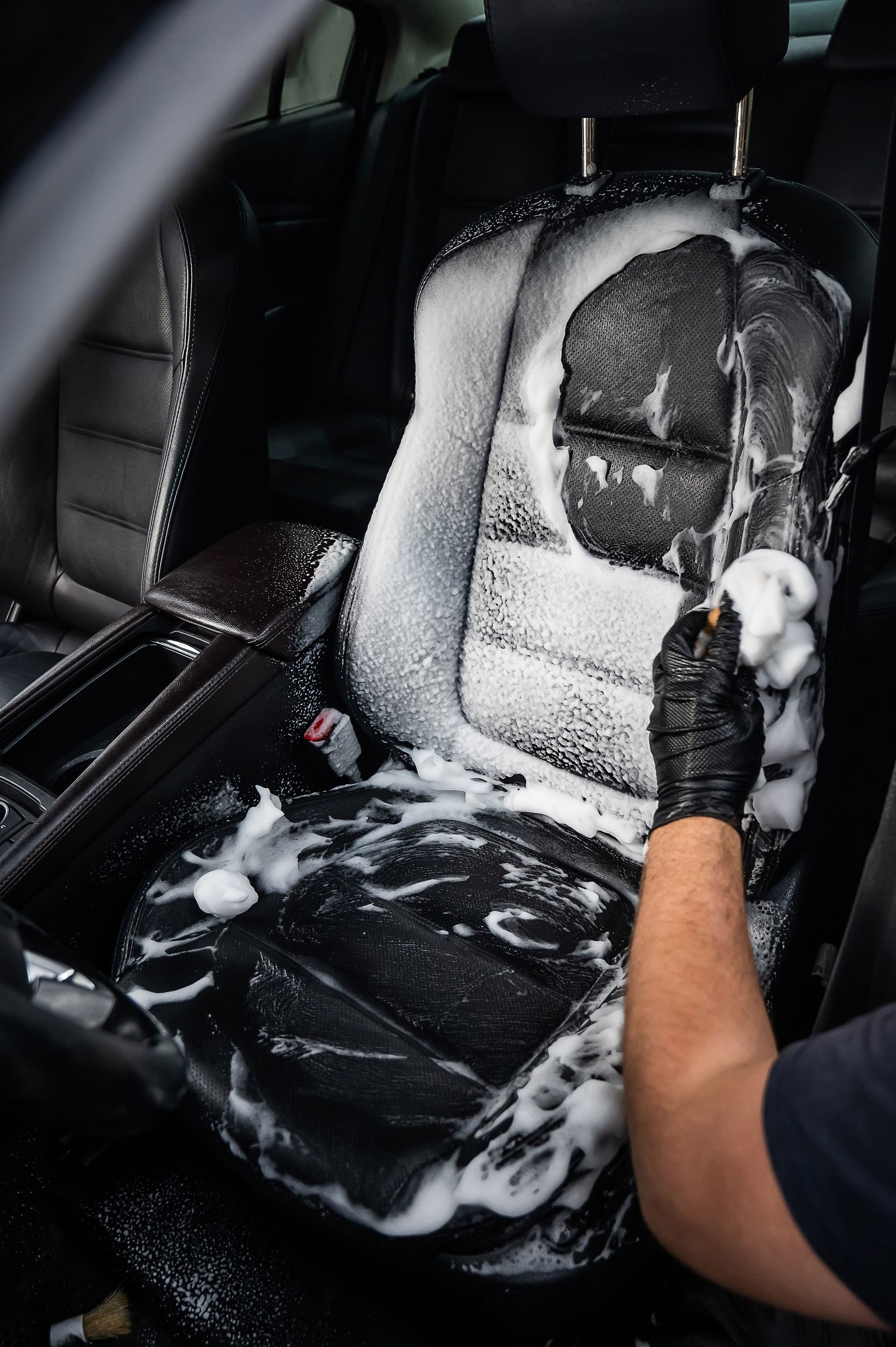 Black-gloved hand scrubbing a car seat with foamy cleaner.