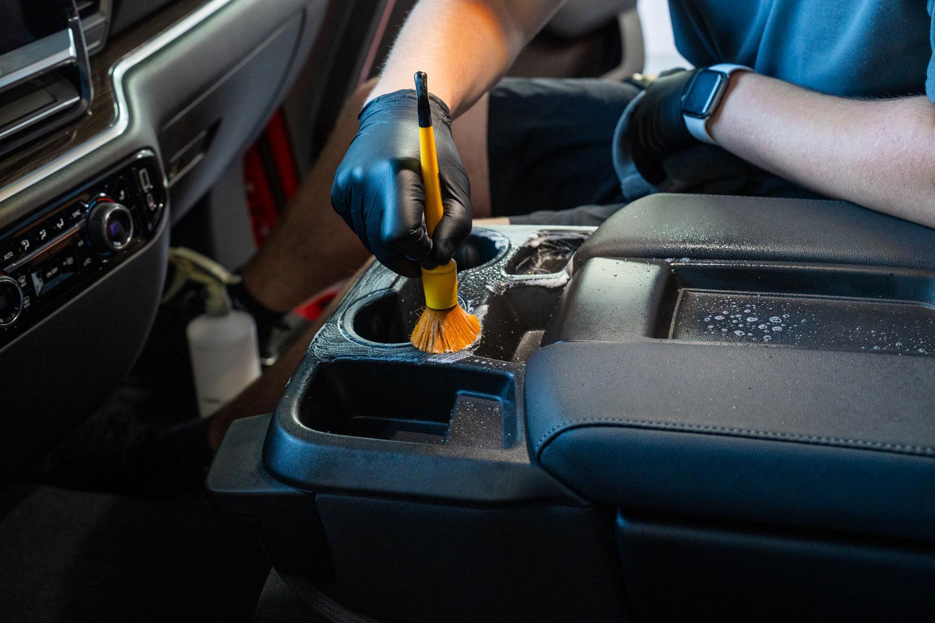 Person wearing black gloves cleaning a car console with a brush.