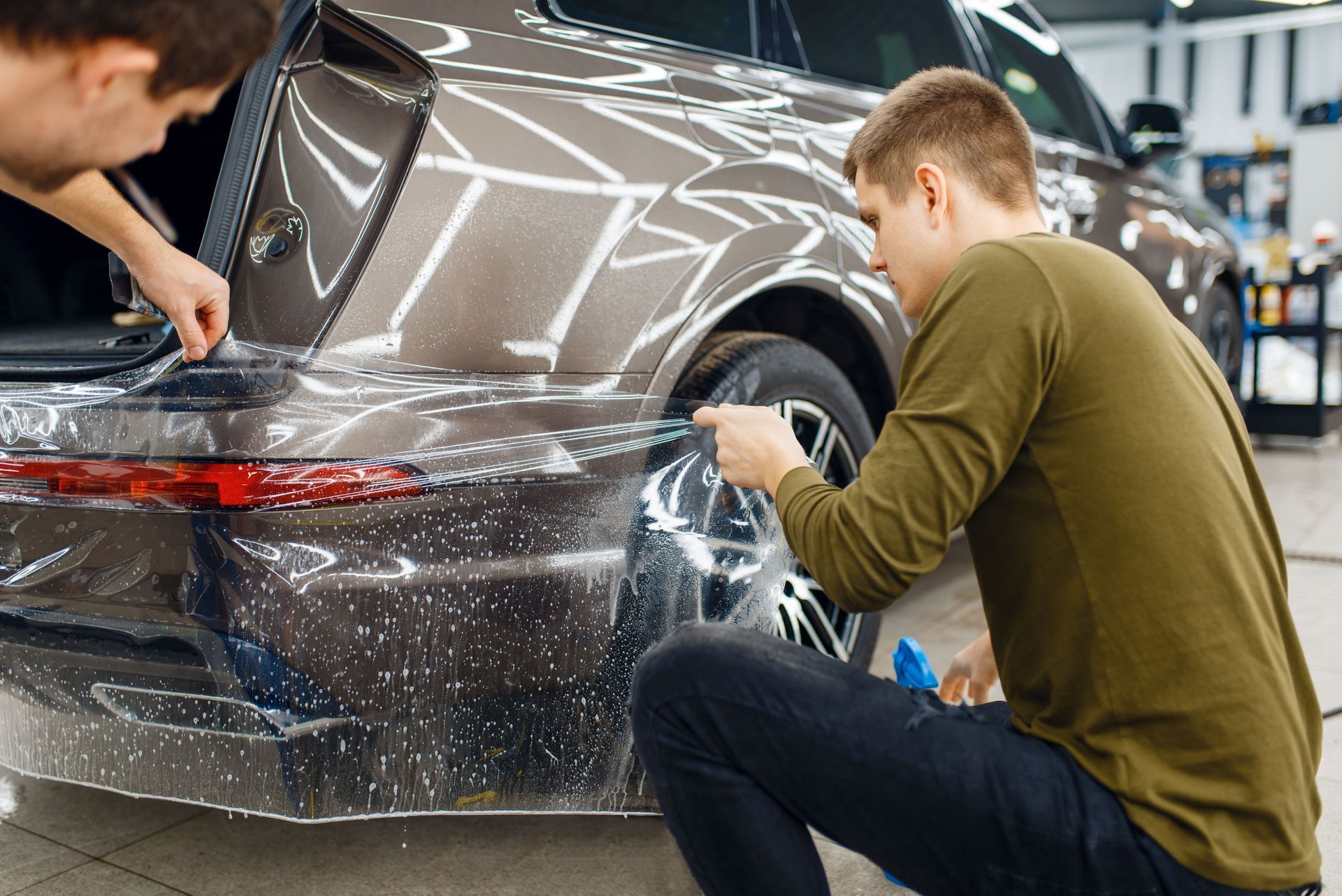 Two people applying protective film to the rear of a brown car in a shop.