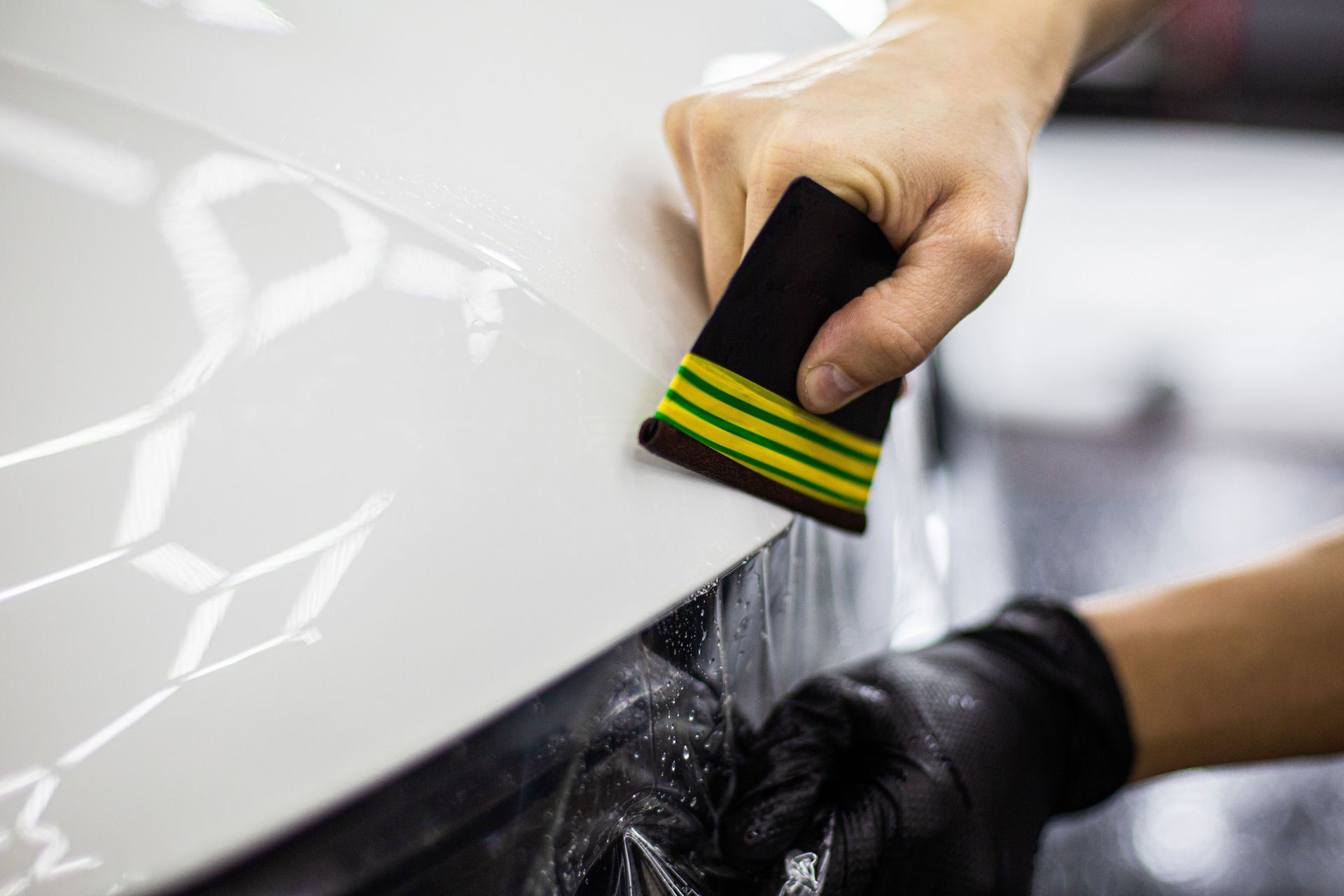 Person applying protective film to a white car's surface with a squeegee. Black gloves, detail shop.