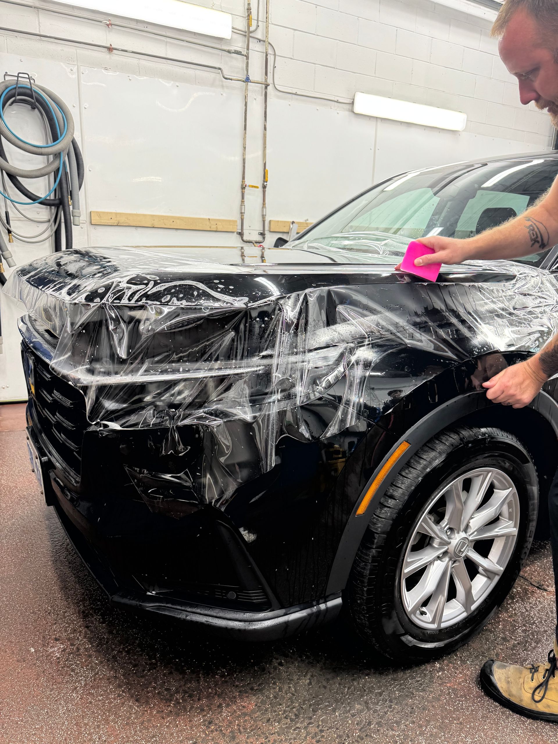 Man applying clear protective film to the front of a black car in a garage.