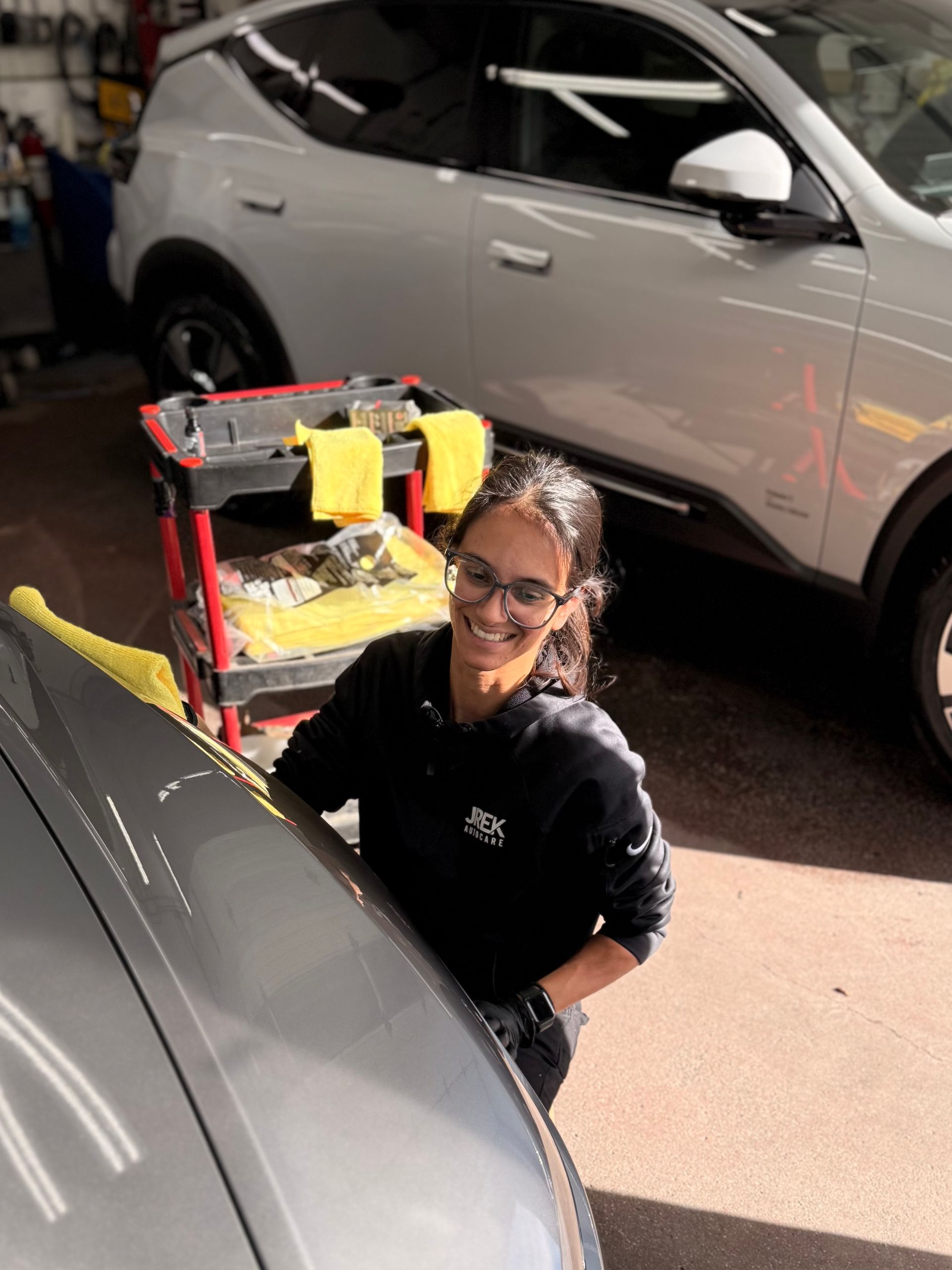 Woman smiles while detailing a grey car in a garage. A detailing cart and a white SUV are in the background.