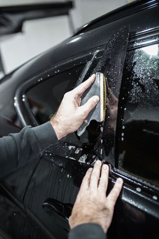 Person applying window tint to a black car window with a squeegee in a professional setting.