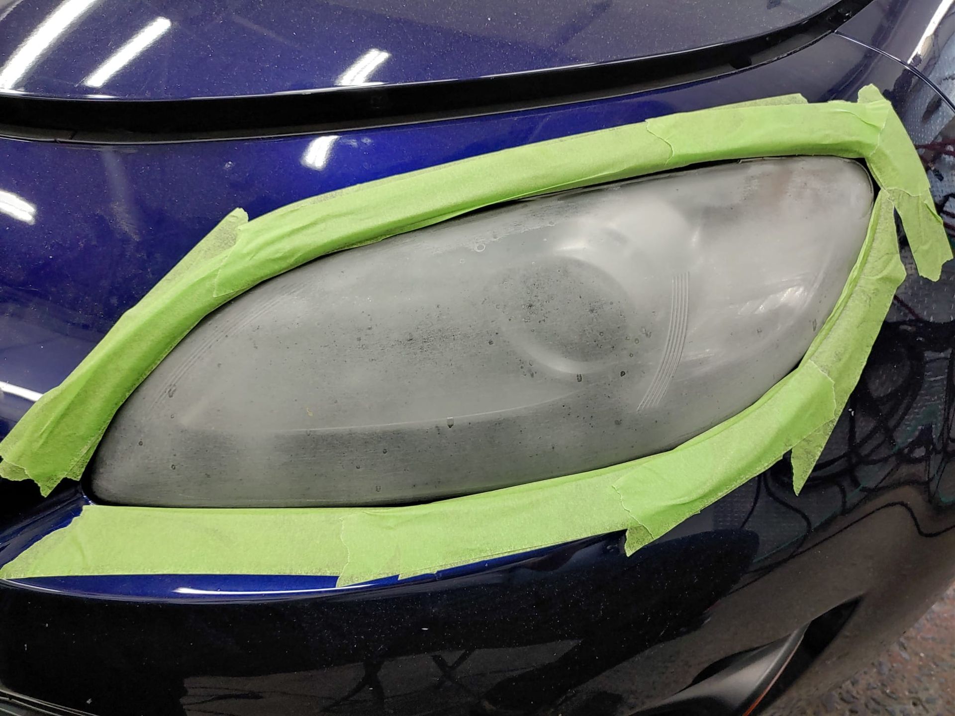 Blue car headlight covered in green tape, being prepared for polishing.