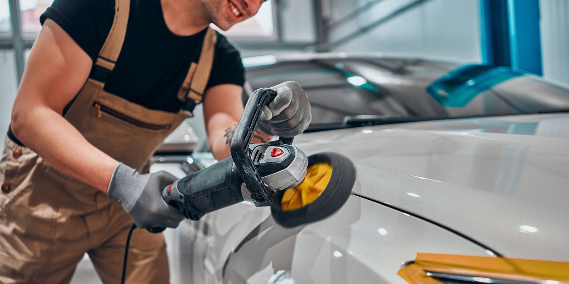 Person in overalls using a polisher on a white car in a garage.