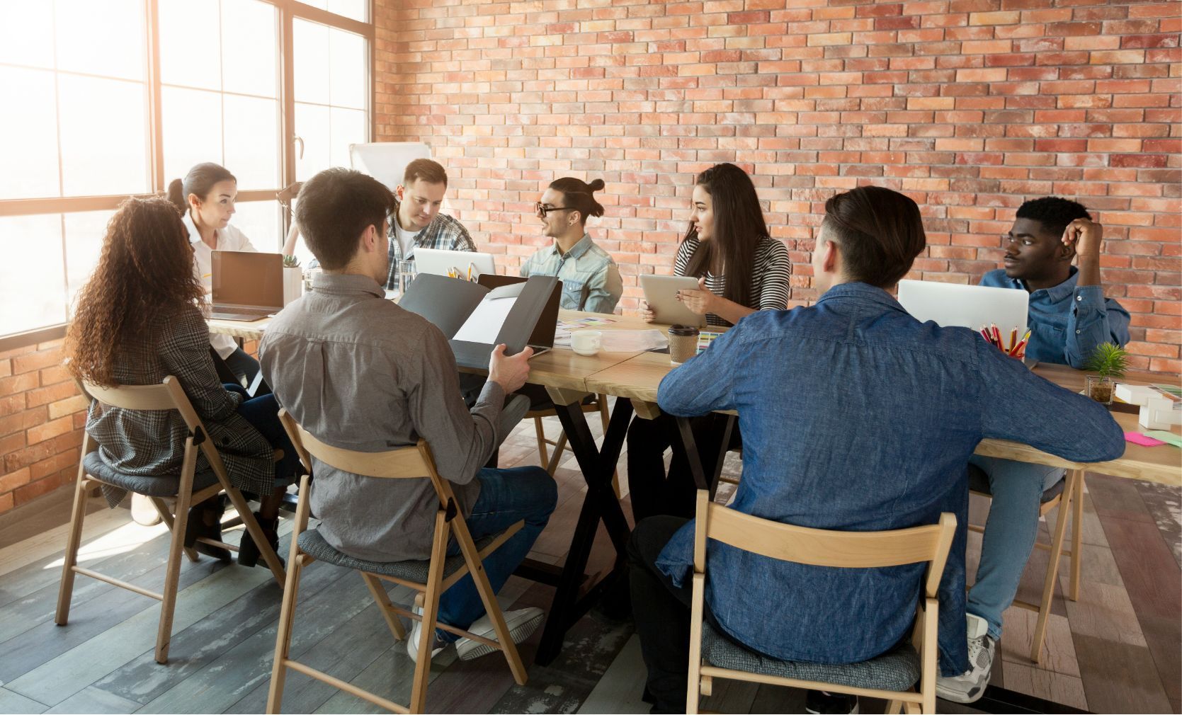 Eine Gruppe von Menschen sitzt in einem Raum mit Backsteinwand um einen Tisch und arbeitet an Laptops und Papieren.