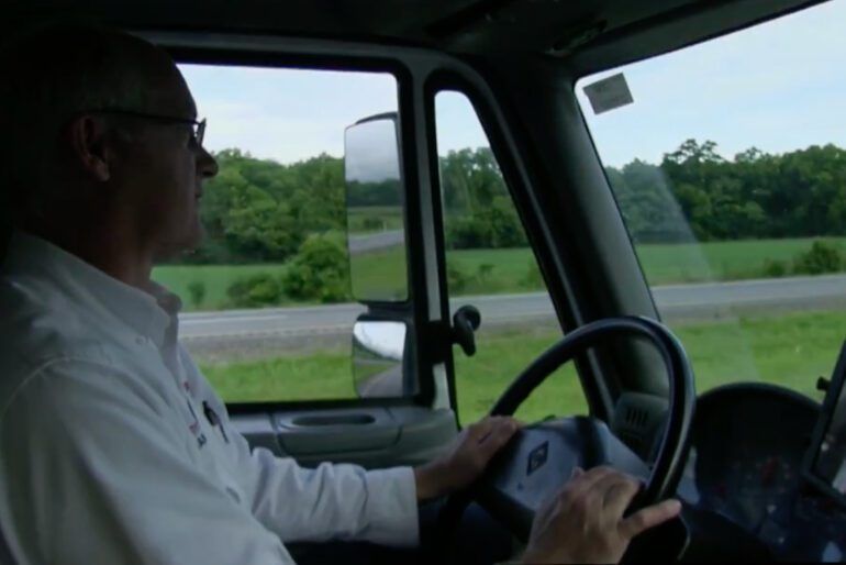 Driver in a truck cab steering along a rural road with green fields outside the window.