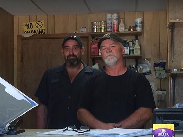 Two men standing behind a counter in a wood-paneled shop, with shelves and a “No Smoking” sign behind them.