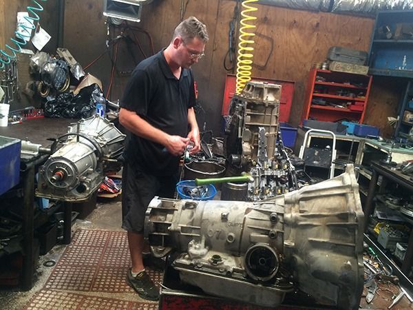 Mechanic working on a large transmission in a workshop, with engine parts and tools around him
