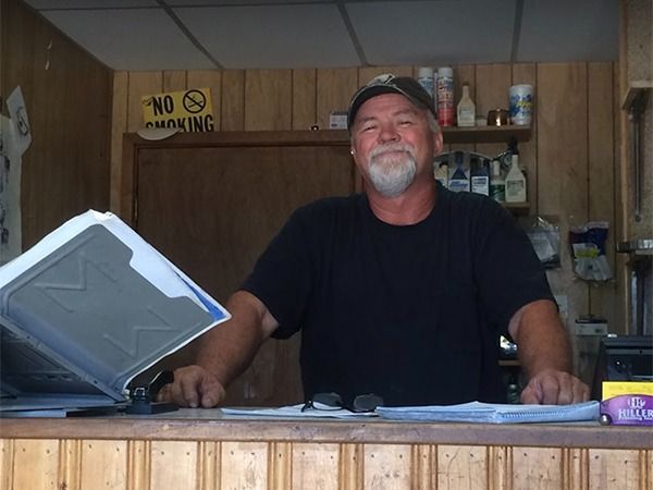Smiling man in a black shirt behind a counter in a small shop, with a cash register open.