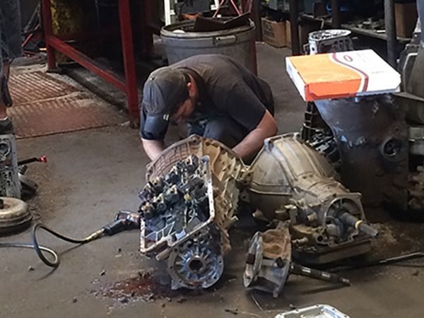 Mechanic crouching over a large engine transmission in a cluttered workshop with tools and parts nearby