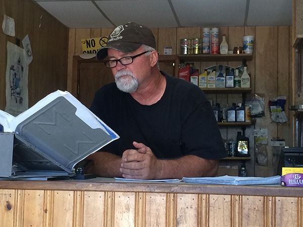 Man reading a large binder behind a counter in a small shop with shelves of goods.
