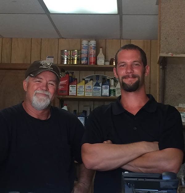 Two men standing in a wood-paneled shop, smiling with shelves of supplies behind them.