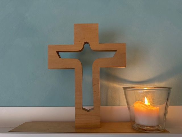 Ein Holzkreuz mit einer ausgeschnittenen Figur steht neben einer brennenden Kerze in einem Glashalter. Es befindet sich vor einer blauen Wand.