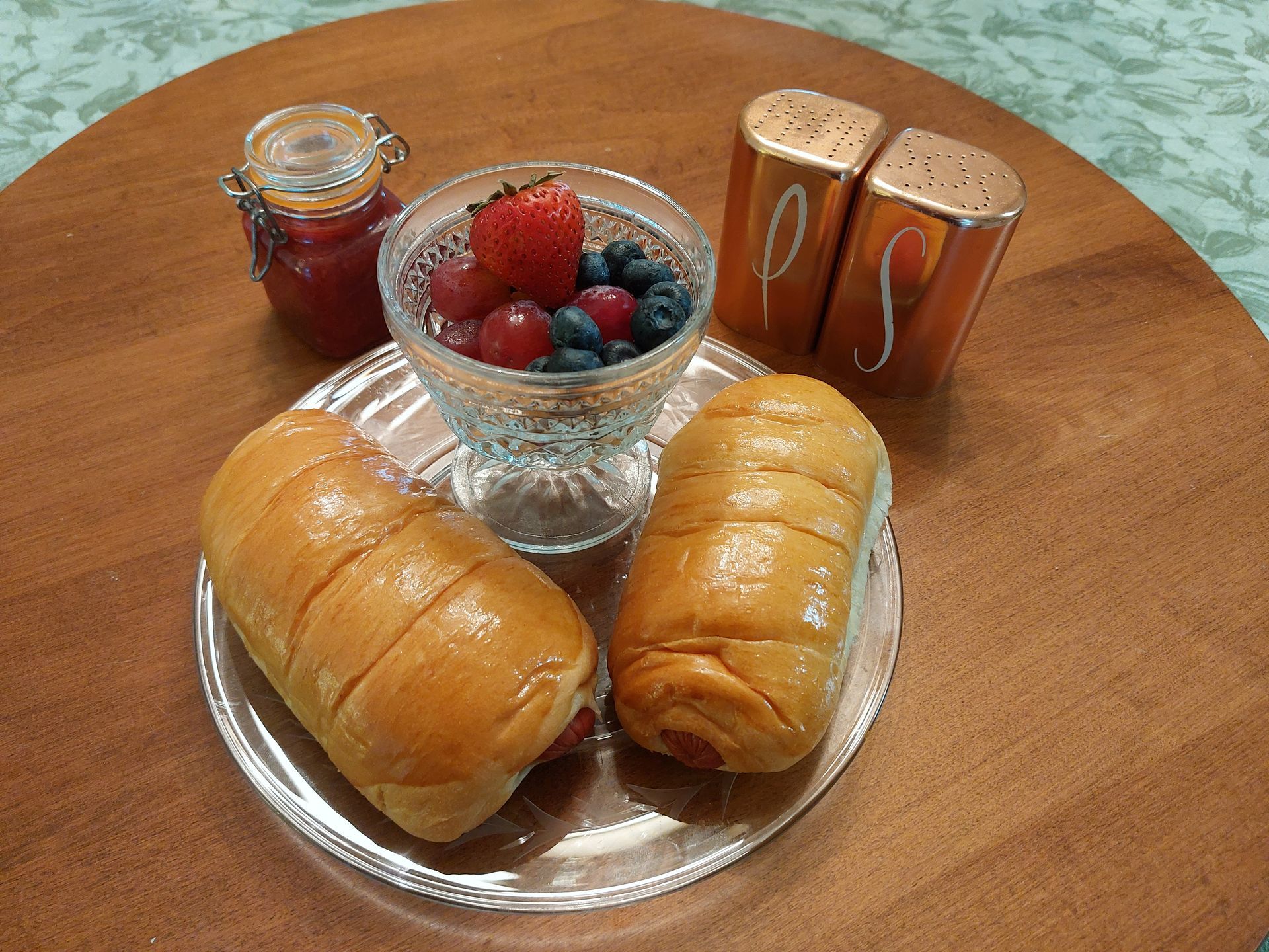 Two hot dogs on a plate next to a bowl of berries and salt and pepper shakers