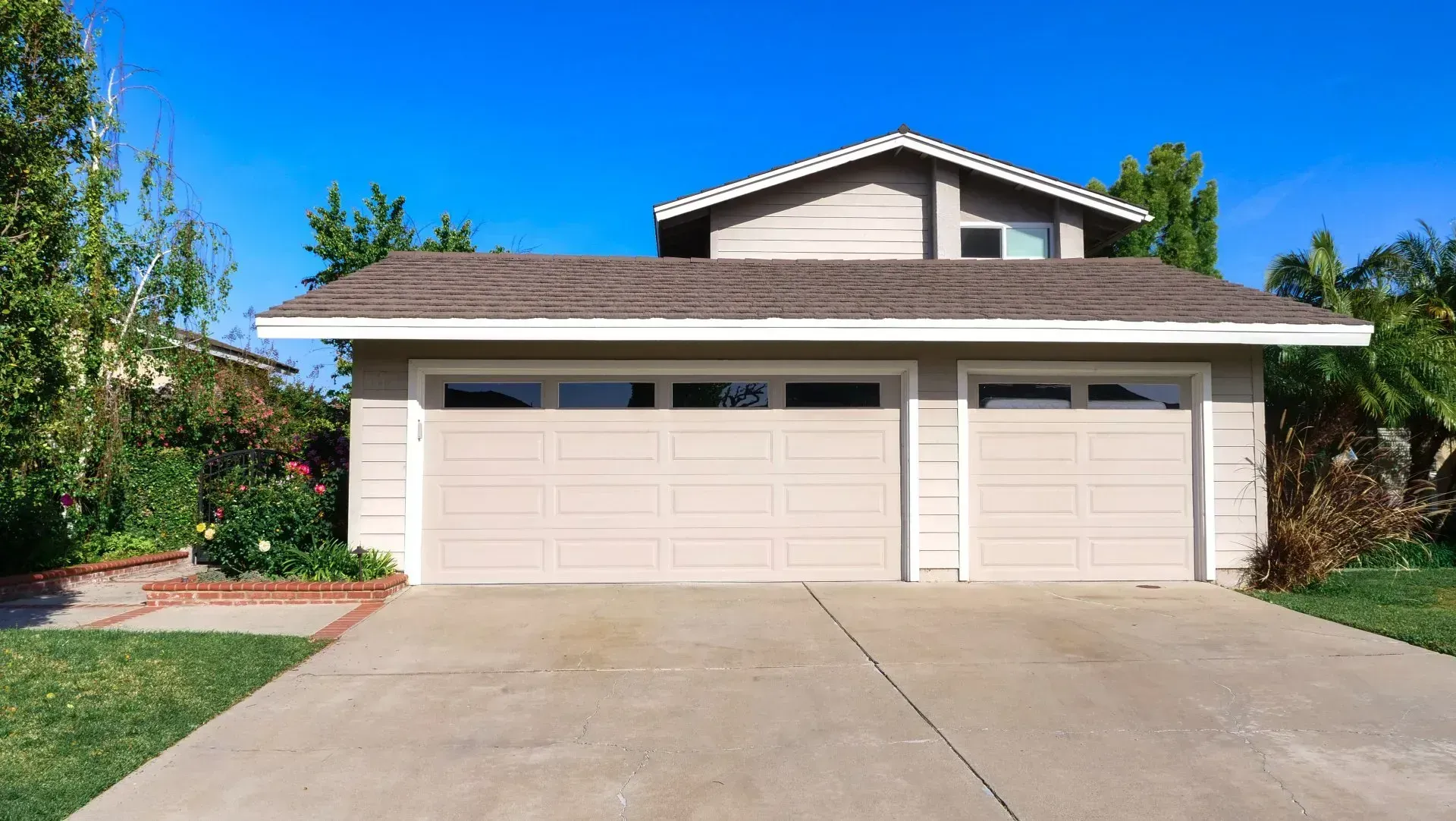Two-car garage with tan doors, brown roof, and a driveway in front of a house against a clear blue sky.