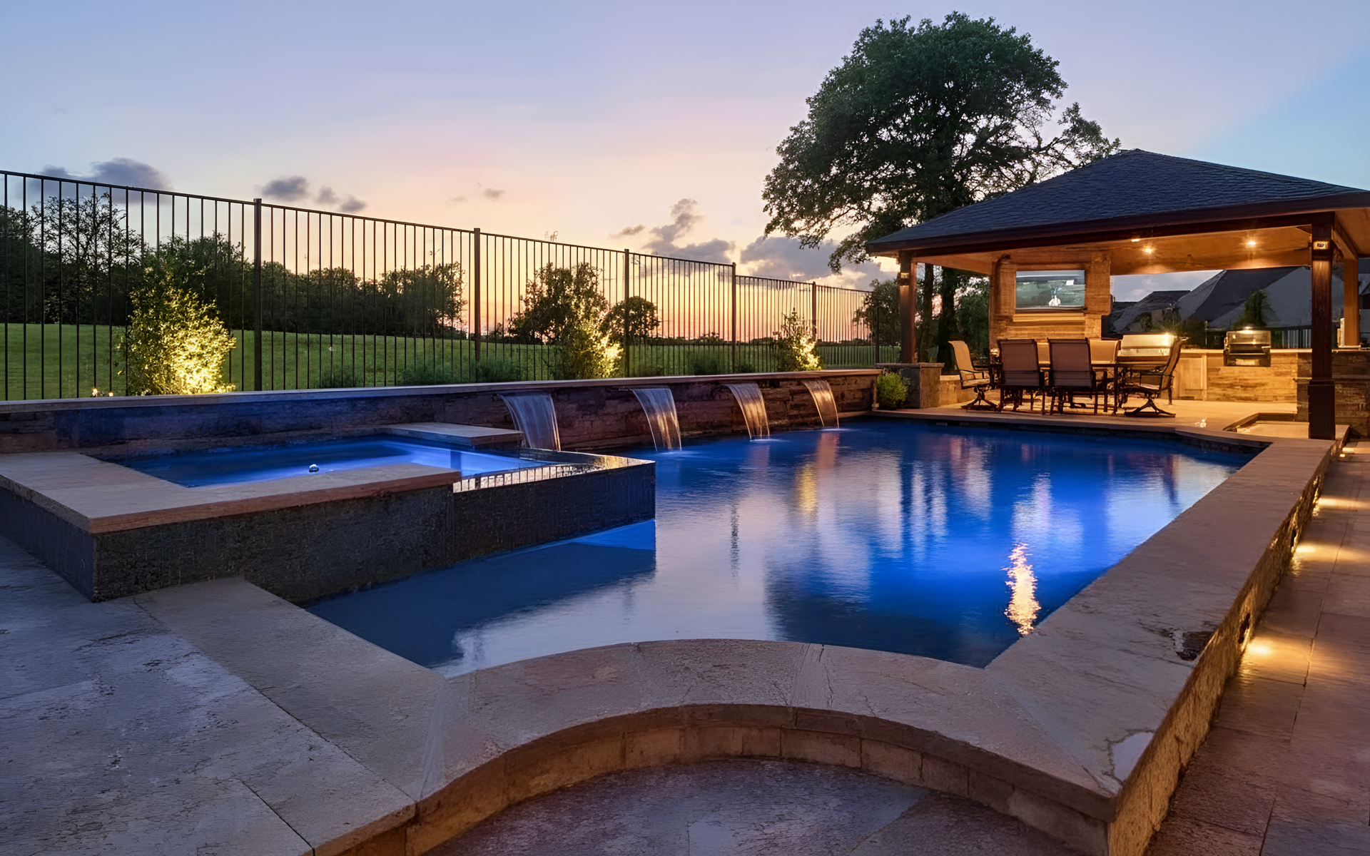 A luxurious pool area at dusk with a gazebo, waterfalls, and illuminated water.