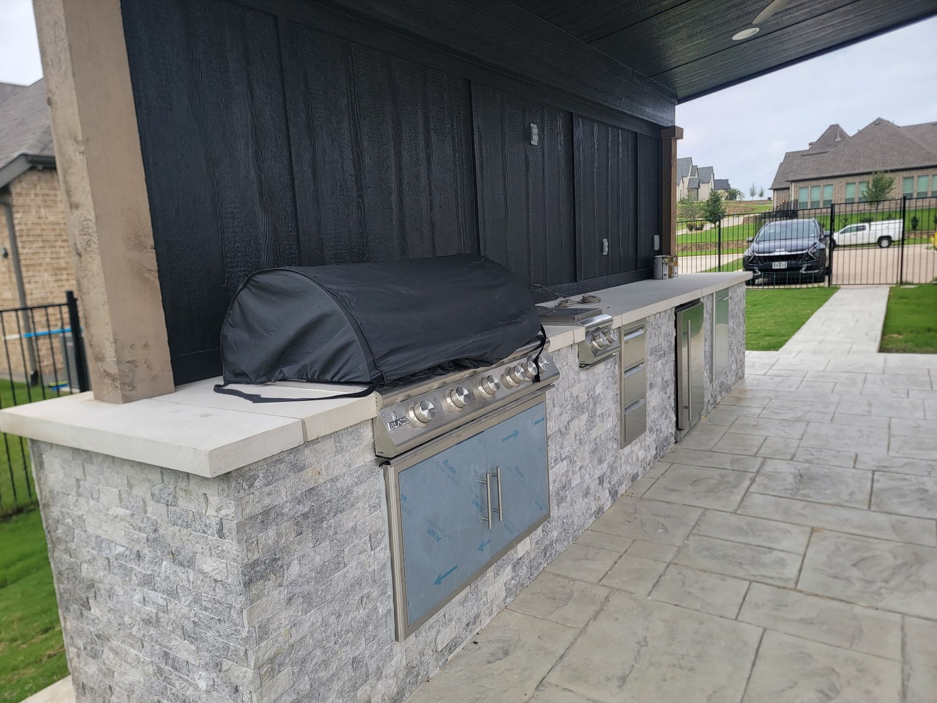 Outdoor stone barbecue grill station beside a paved patio and driveway under a covered structure
