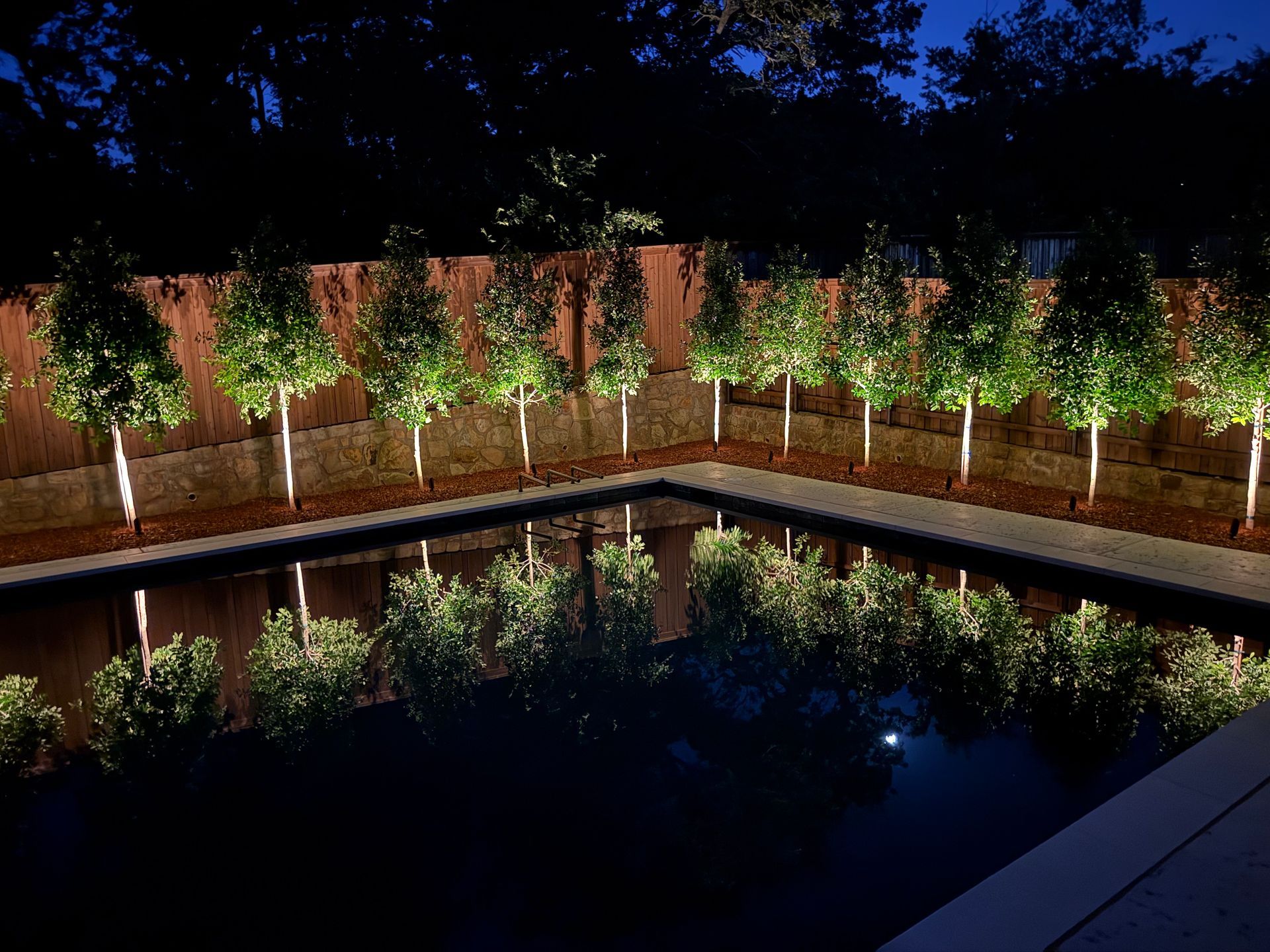 Nighttime courtyard with lit trees and a reflecting pool beside a brick wall