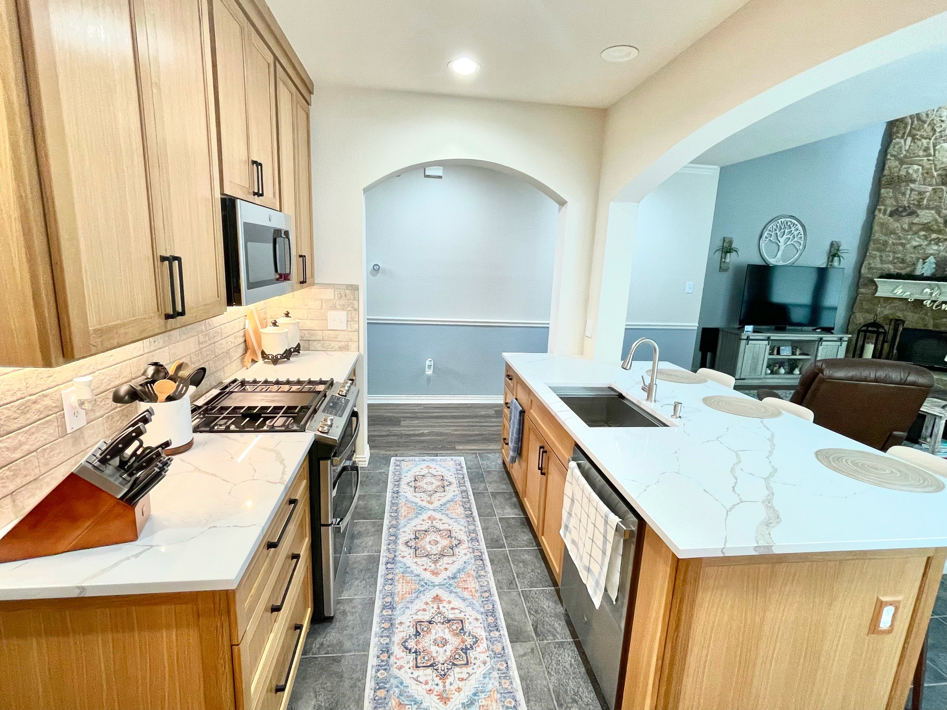 Kitchen with light wood cabinets, white countertops, island, and rug. Archway leads to another room.