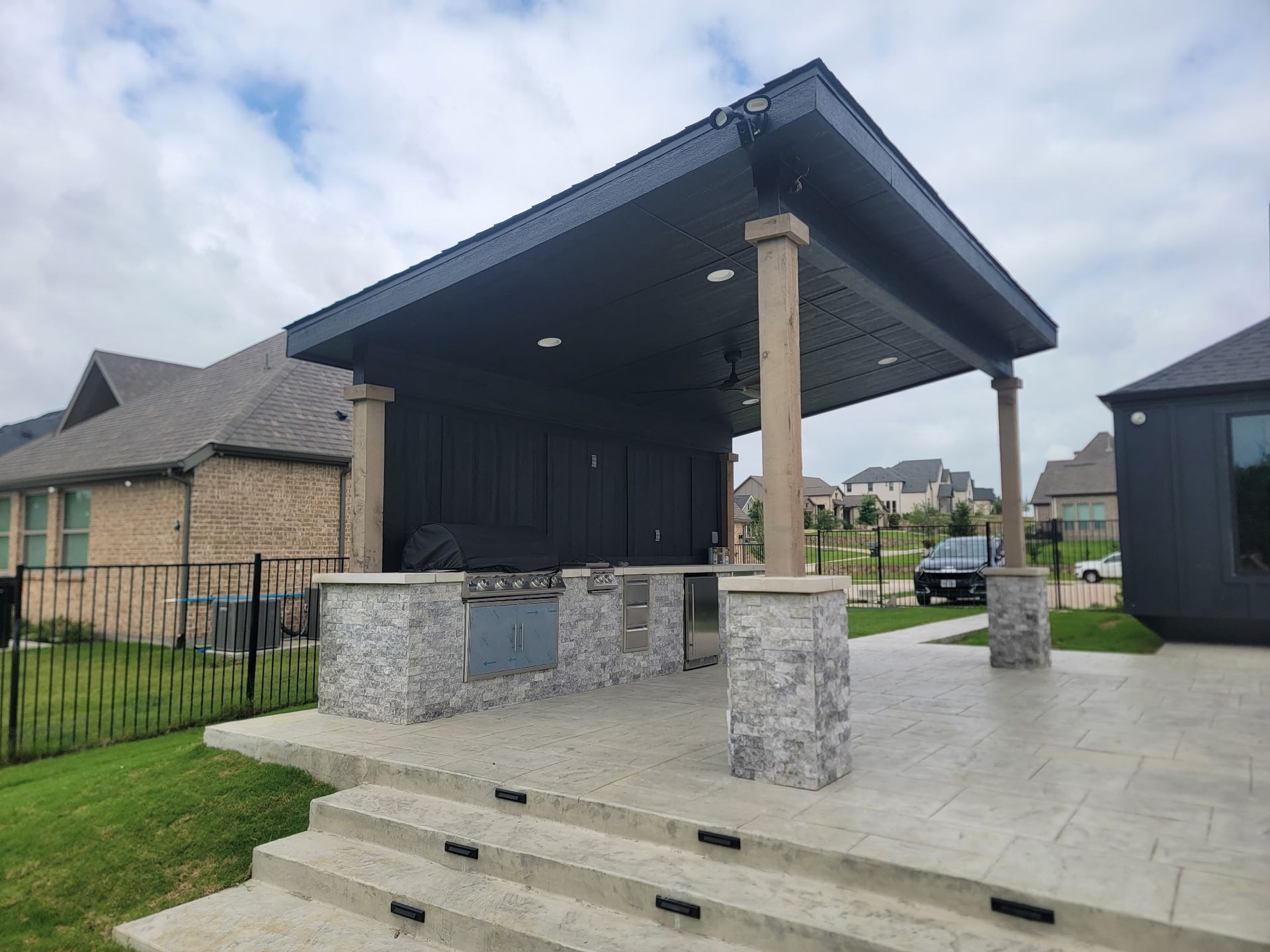 Covered outdoor patio with stone columns, tiled floor, and a small stage beside houses and parked cars.