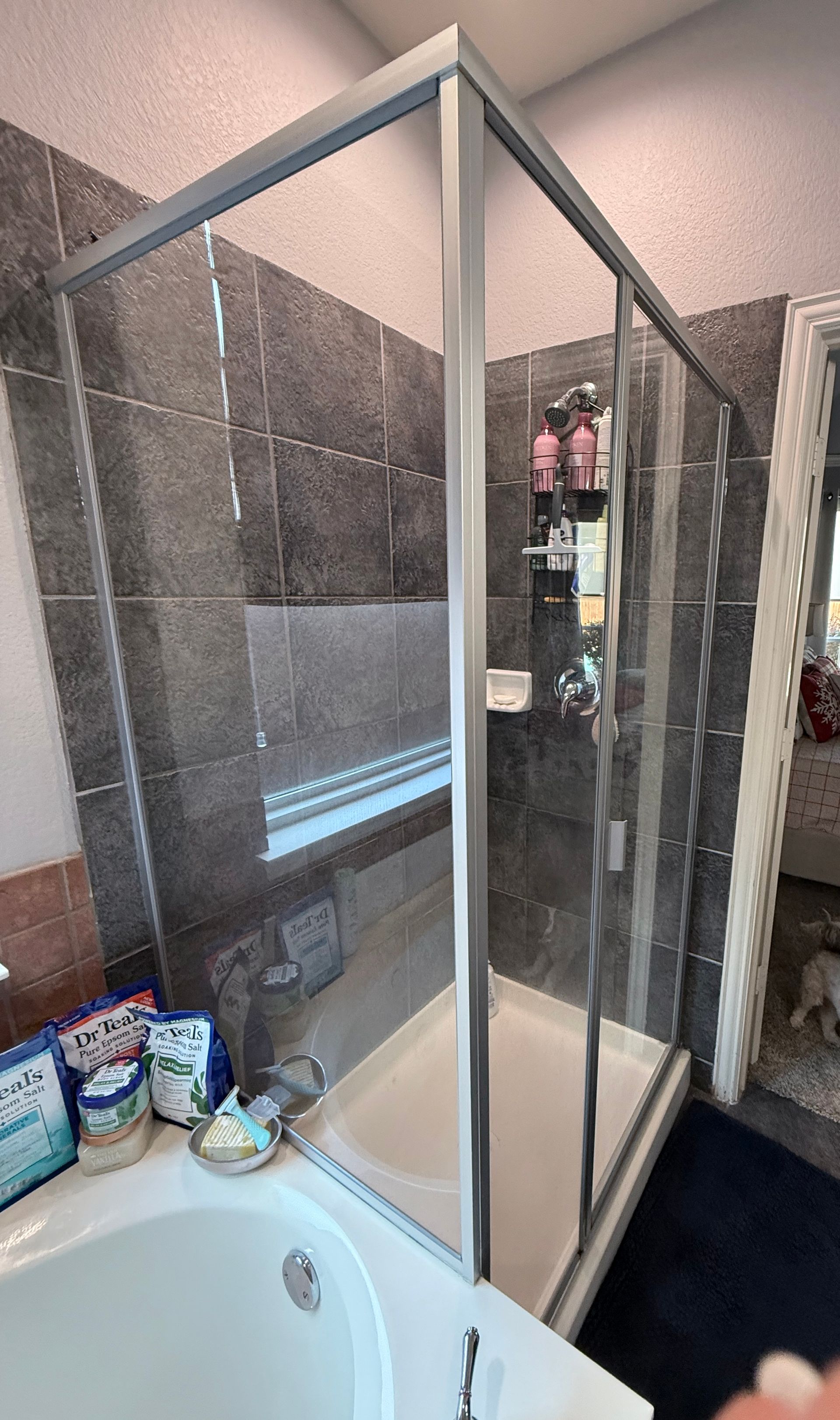 Corner shower with glass doors in a bathroom, next to a bathtub and toiletries on the edge.