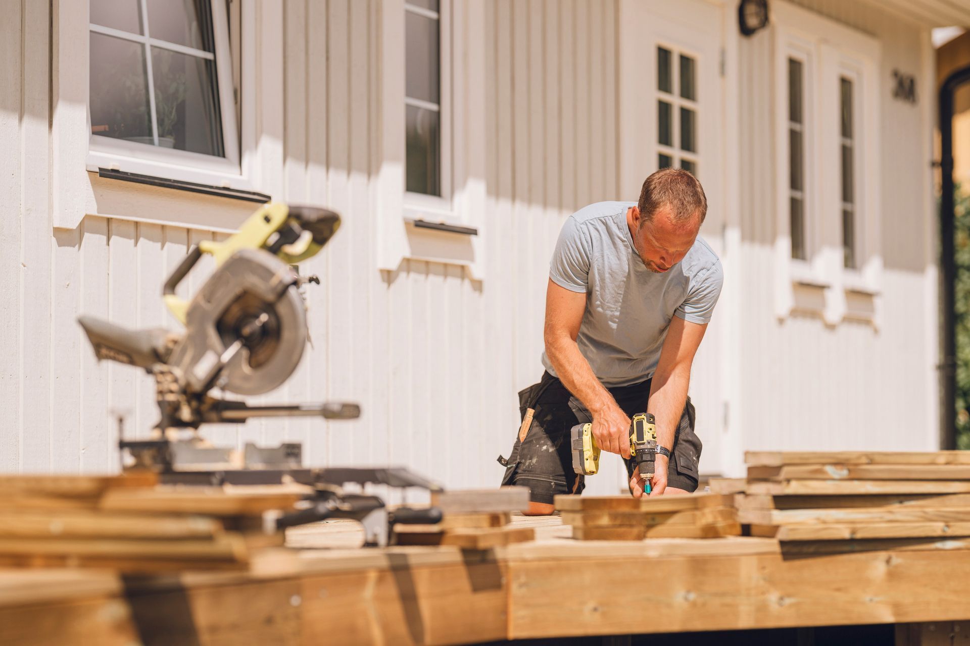 Man using a power drill on a wooden deck. Man using a drill on a wooden deck in front of a white house.