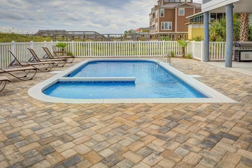 Rectangular in-ground pool surrounded by brick patio. White fence and beach in the background, near a house.
