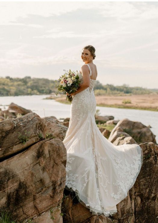 A bride in a wedding dress is standing on a rock near a lake holding a bouquet of flowers.