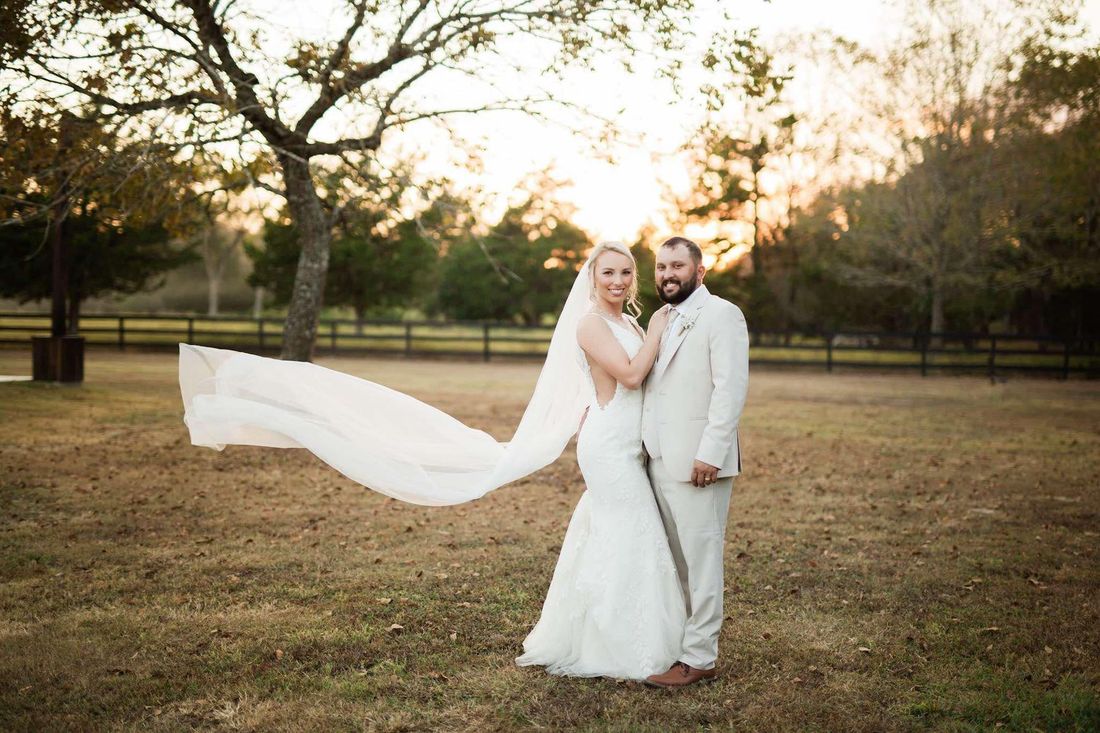 A bride and groom are posing for a picture in a field with a veil blowing in the wind.