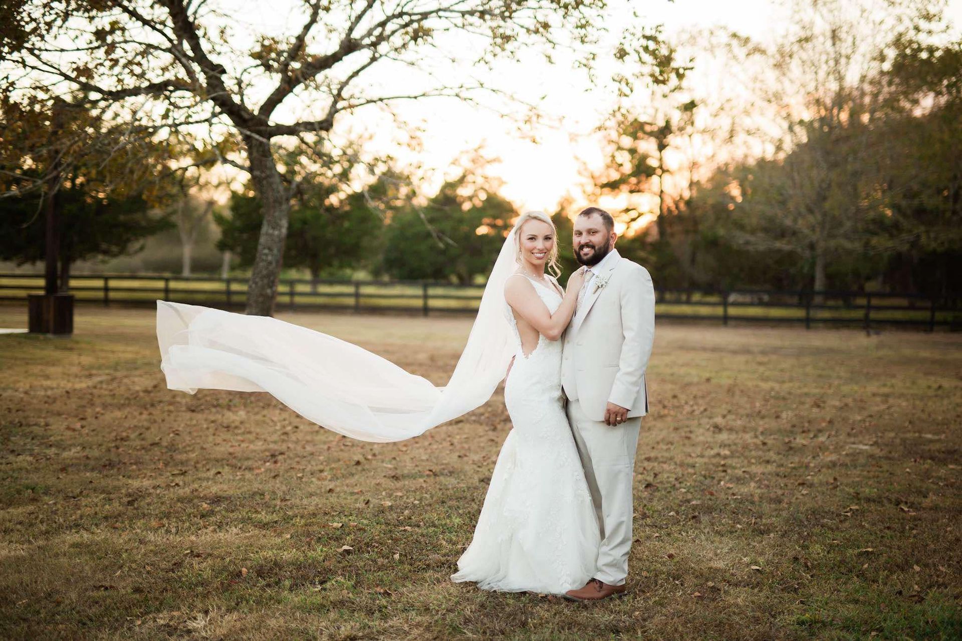 A bride and groom are posing for a picture in a field with a veil blowing in the wind.
