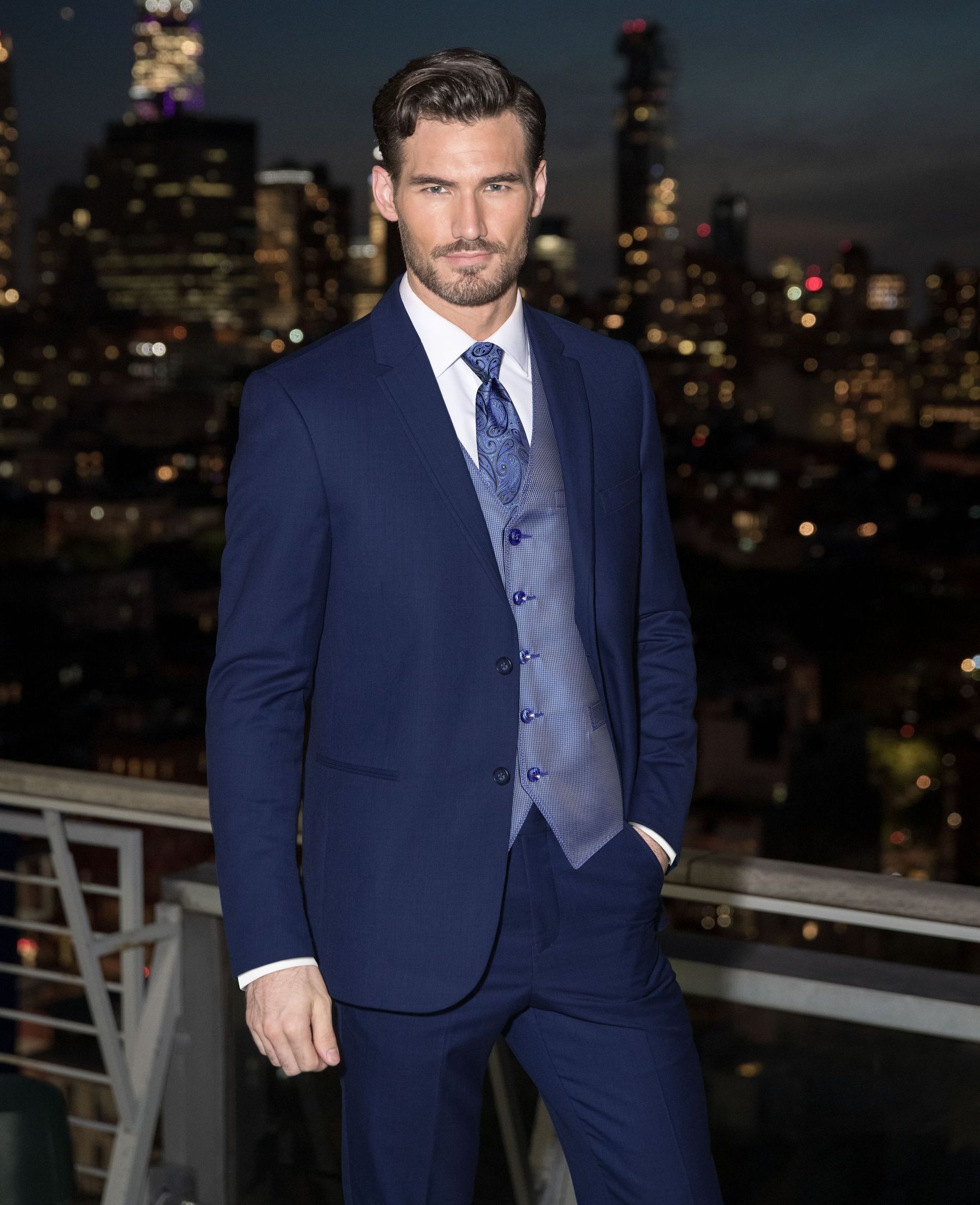 A man in a black suit and yellow tie is standing in front of a stone wall.