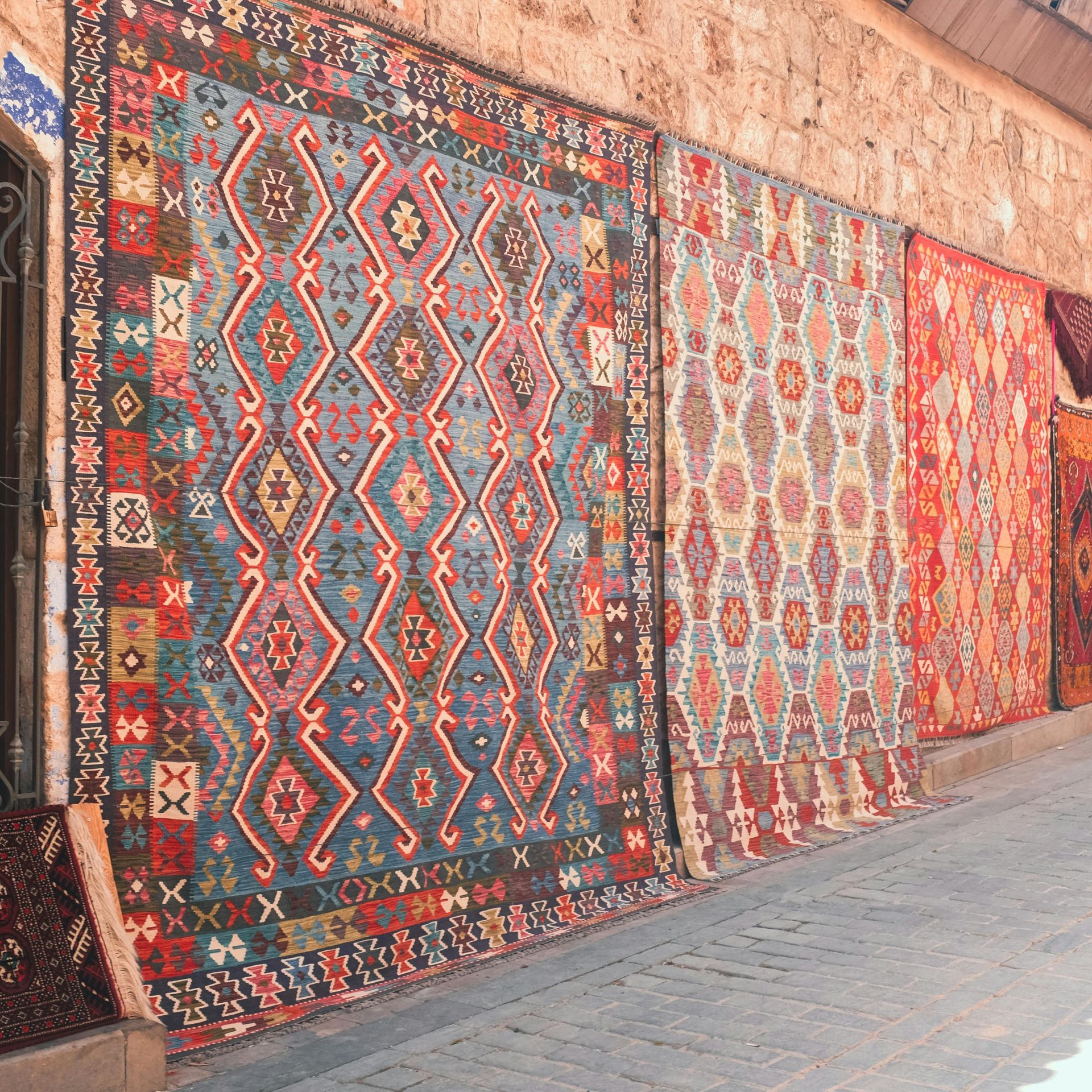 Several colourful, patterned rugs hang on a stone wall along a stone-paved walkway.