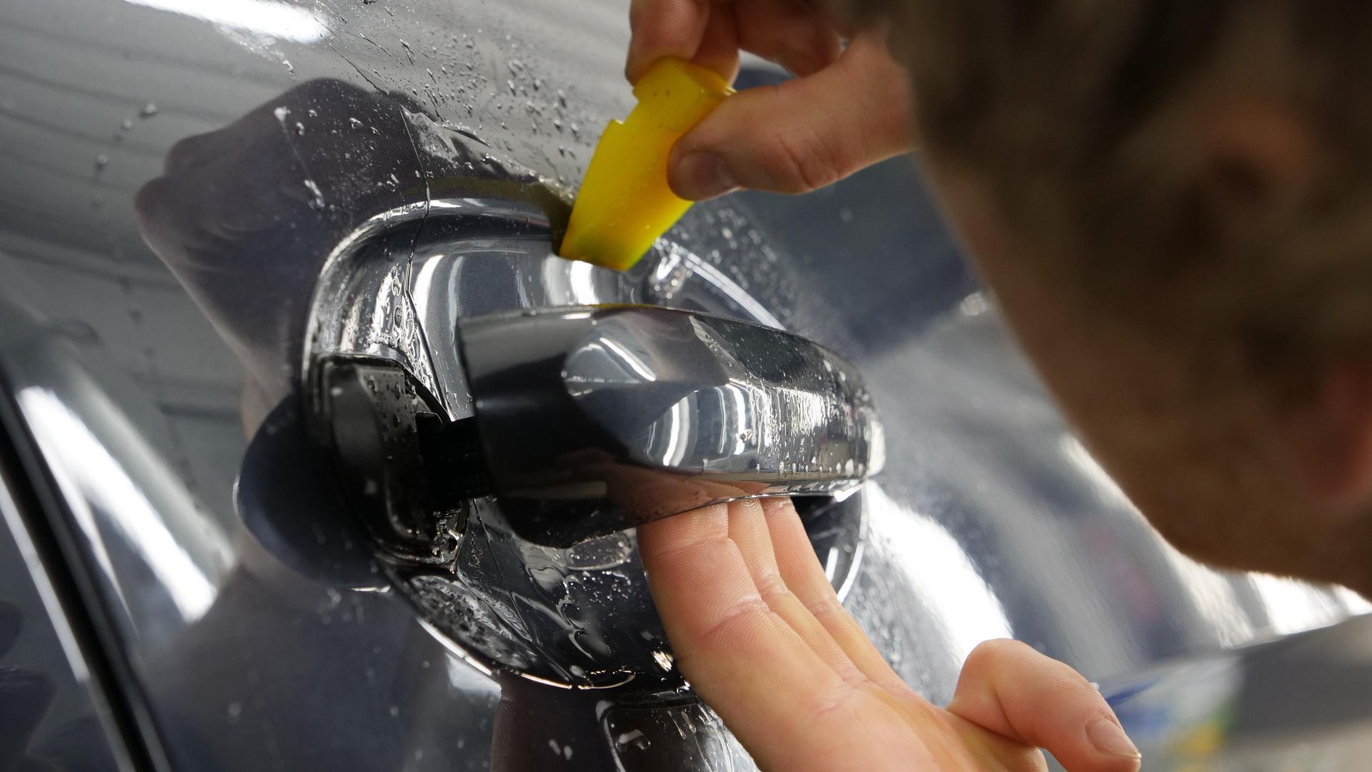Person applying protective film to a car door handle with a yellow squeegee.