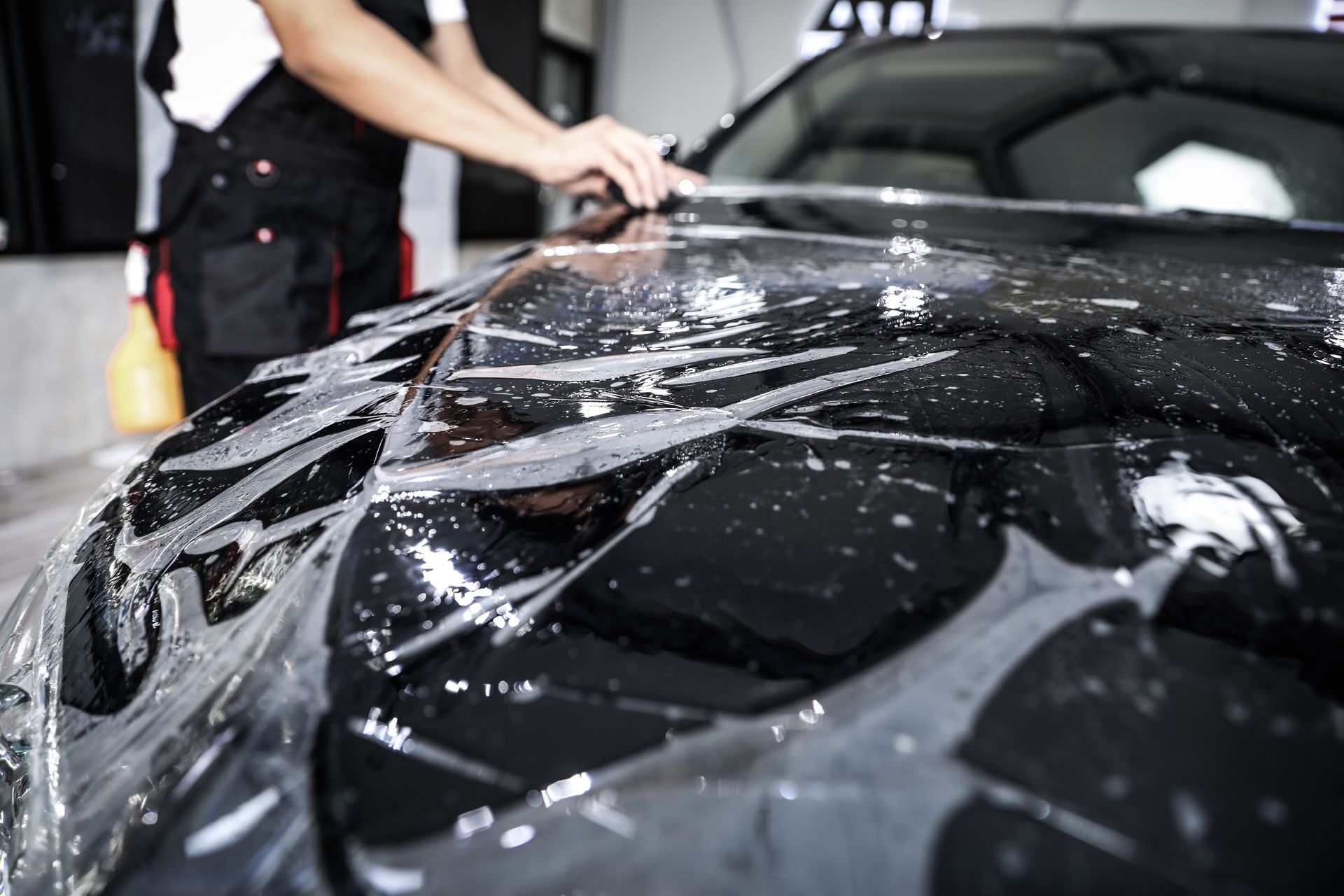 A man is covering the hood of a black car with plastic wrap.