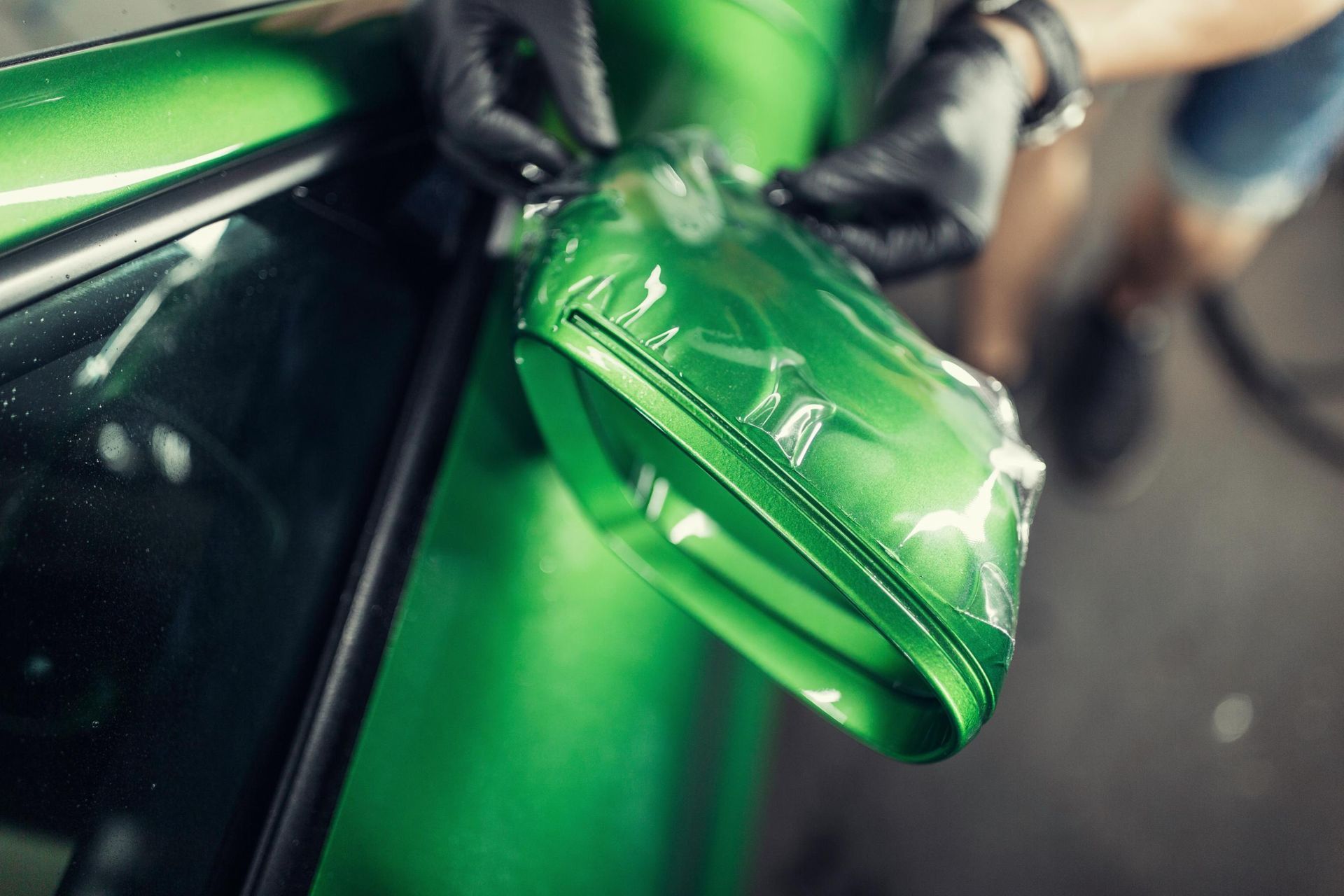 A gloved hand applies a transparent protective film to a bright green car side mirror.