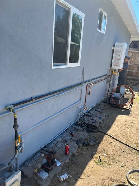 A person installs a wall-mounted water heater on the exterior of a grey house, with gas and water piping running along it.