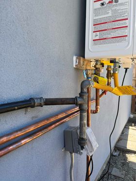A wall-mounted tankless water heater with copper water lines and a black iron gas pipe connection on a gray exterior wall.