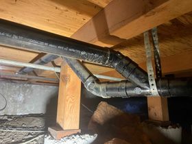 Black ABS plumbing pipes joined by a Y-fitting and supported by metal strapping under a wooden floor structure.
