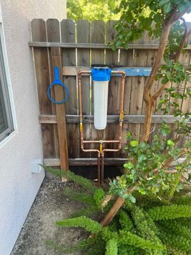 A whole-house water filter housing mounted to a wooden fence with copper plumbing and a blue plastic filter wrench.