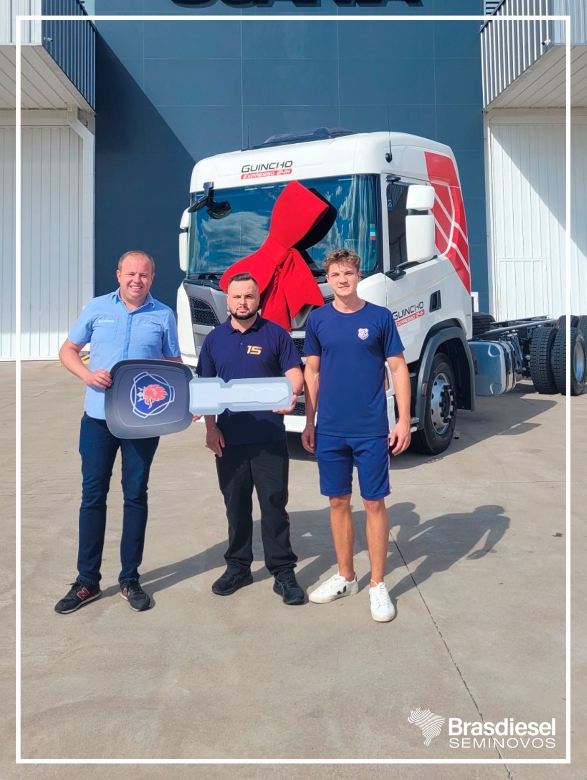 Three men stand in front of a white truck in a warehouse, holding a plaque and a box.