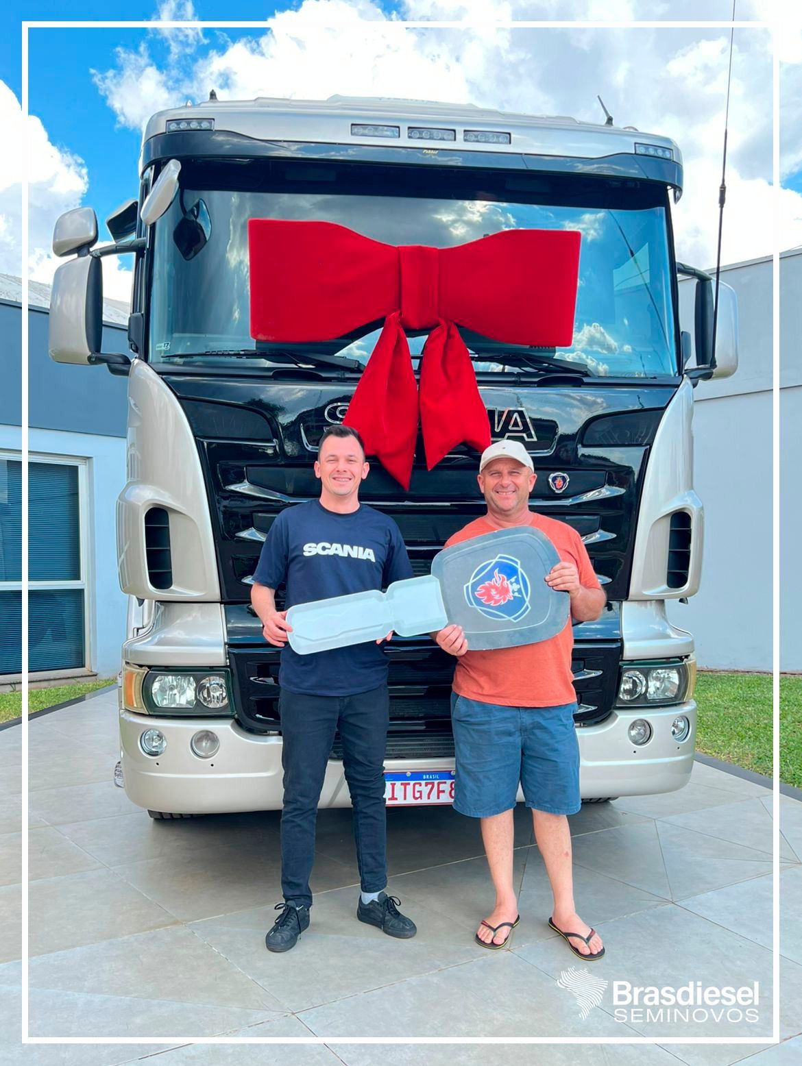 Two men stand in front of a large truck with a big red bow, holding a gift box.