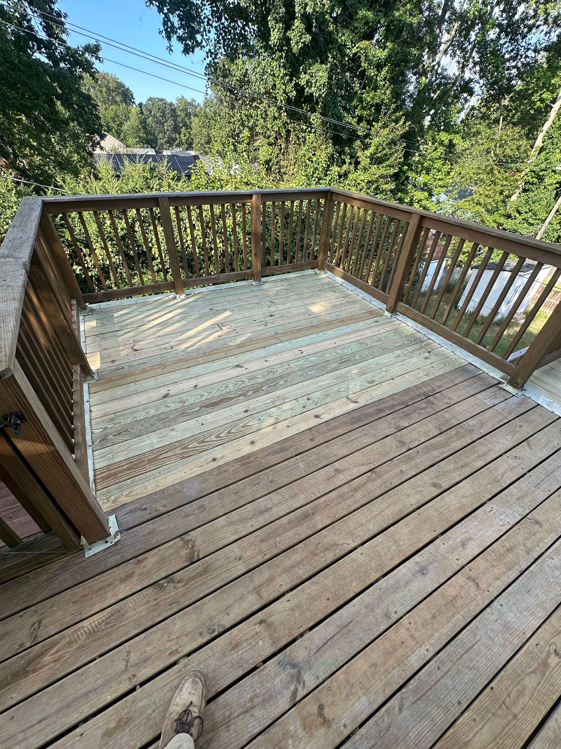 Wooden deck with railing surrounded by lush greenery on a sunny day.
